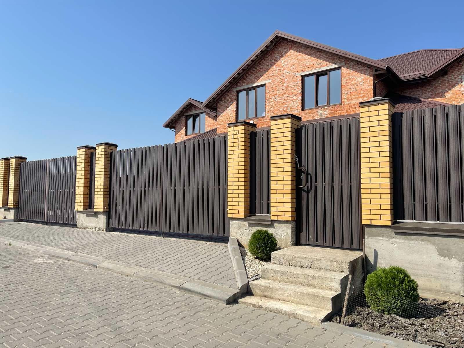 Nowoczesne ogrodzenie z brązowych paneli i żółtej cegły, z bramą wejściową i schodami, na tle domu z czerwonej cegły pod błękitnym niebem.