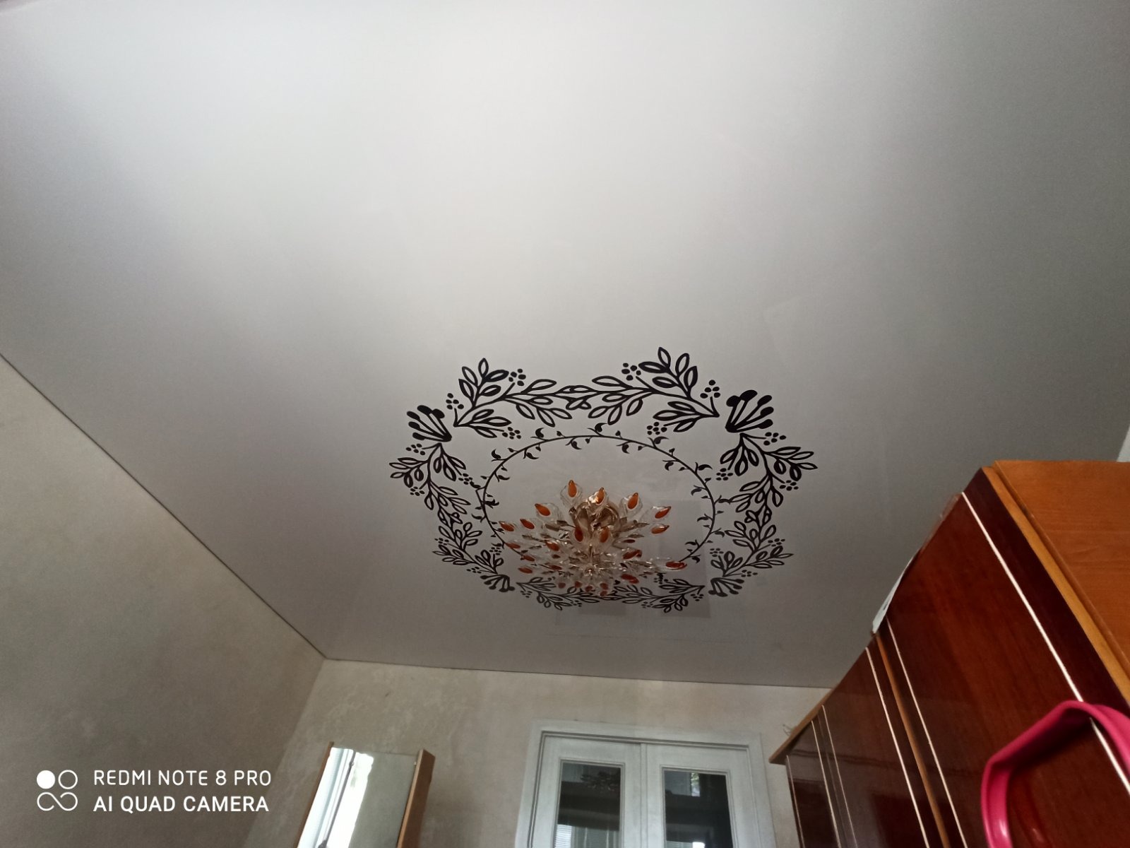 Biały sufit z dekoracyjnym, okrągłym wzorem roślinnym wokół żyrandola z pomarańczowymi elementami. Widoczne fragmenty ścian, okna i szafy.
