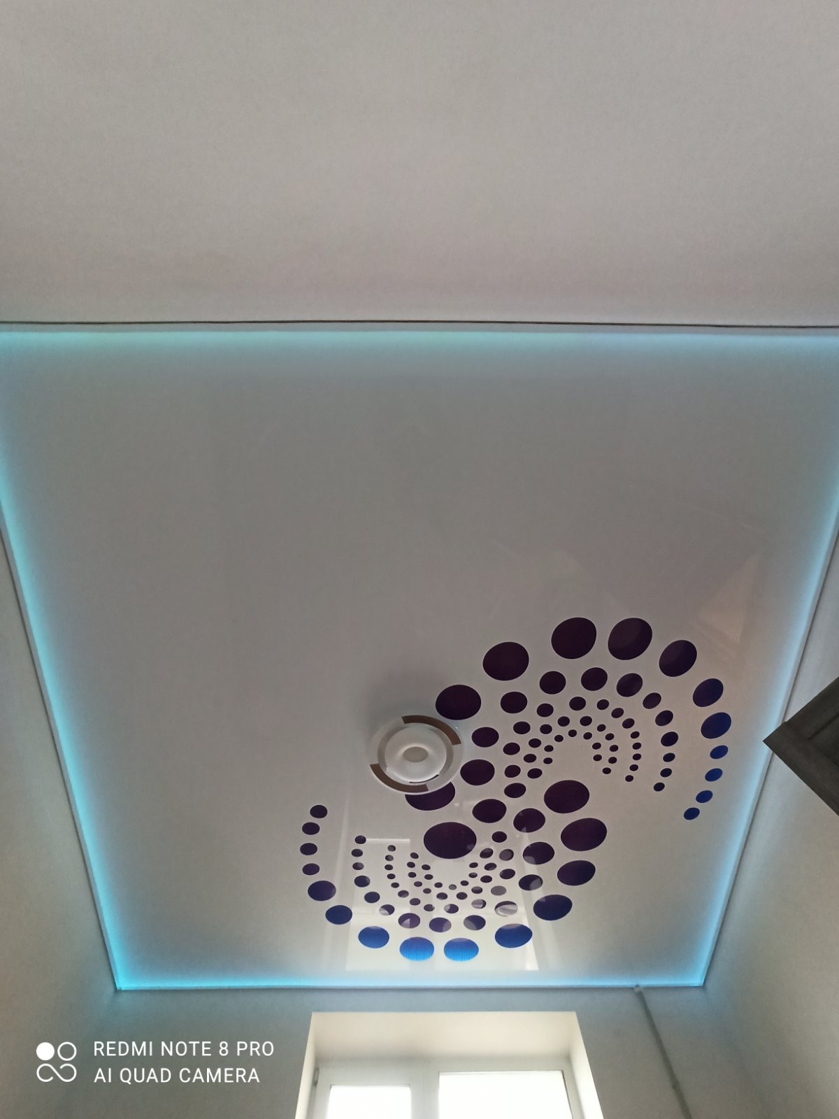 Biały sufit napinany z podświetleniem LED i wzorem z fioletowych kropek, centralnie umieszczony okrągły element oświetleniowy.