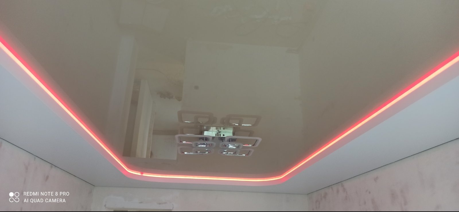 Biały, błyszczący sufit napinany z dekoracyjnym oświetleniem LED wzdłuż krawędzi. Na suficie widoczne odbicie lampy. Ściany w stanie surowym, przed wykończeniem.