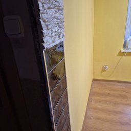 F.w.b.Kreft Firma Wykończeniowo Budowlana Mateusz Kreft - Fragment ściany z dekoracyjnym kamieniem i połyskującymi, brązowymi płytkami w trakcie montażu, widoczne żółte ściany i fragment podłogi w pomieszczeniu.