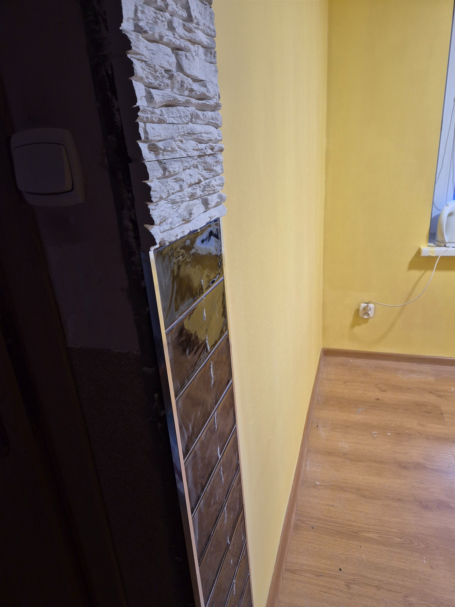 Fragment ściany z dekoracyjnym kamieniem i połyskującymi, brązowymi płytkami w trakcie montażu, widoczne żółte ściany i fragment podłogi w pomieszczeniu.
