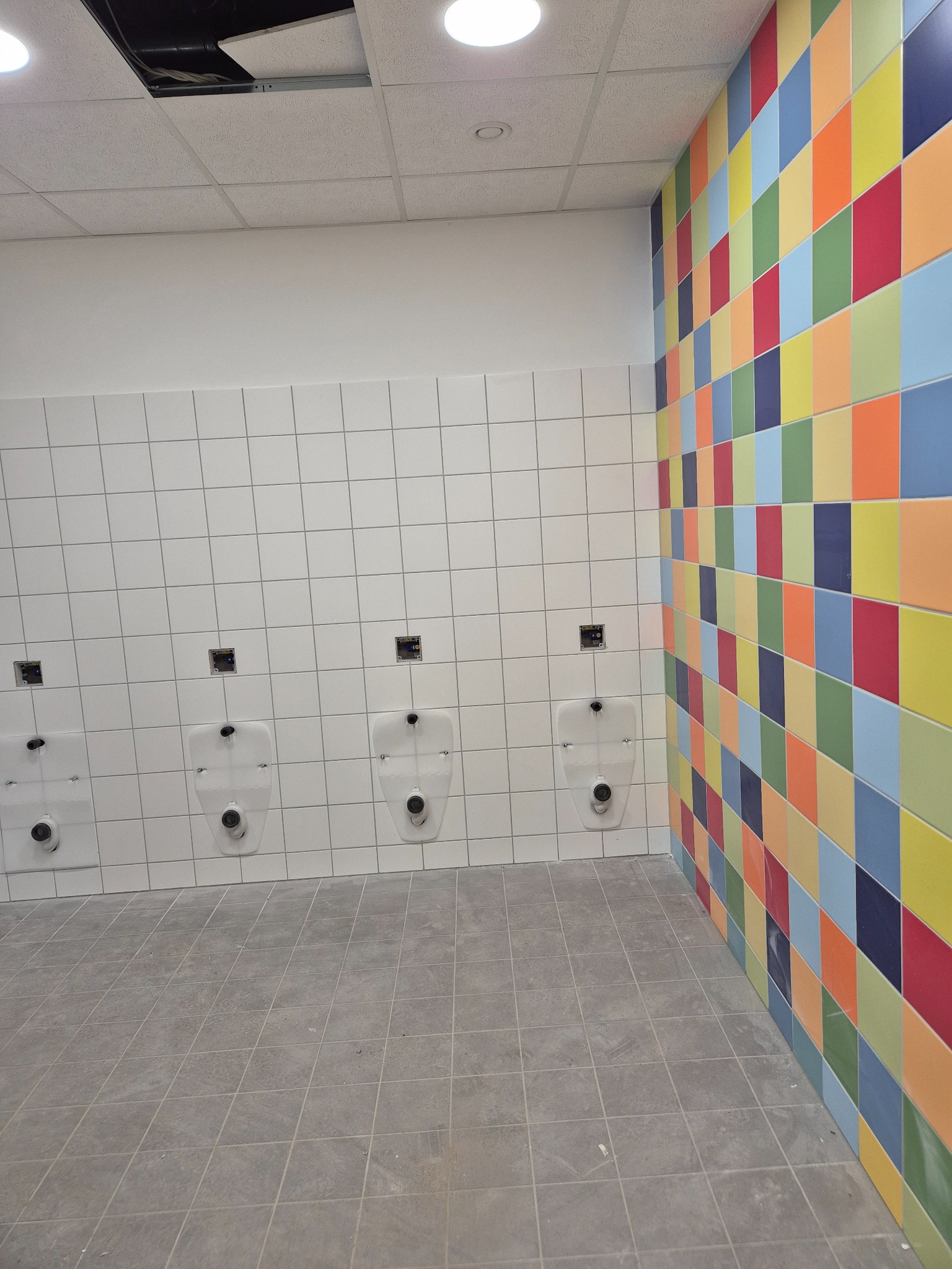 Wnętrze łazienki publicznej z białymi płytkami i rzędem pisuarów. Jedna ściana wyłożona kolorowymi, kwadratowymi płytkami. Sufit kasetonowy z widoczną instalacją.