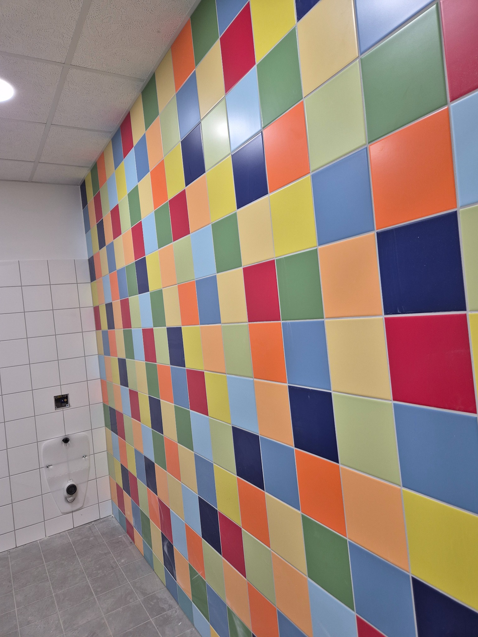 Kolorowa mozaika z kwadratowych płytek w łazience, z widocznym pisuarze na ścianie z białych płytek. Kontrastowe, intensywne barwy płytek tworzą unikalny wzór.