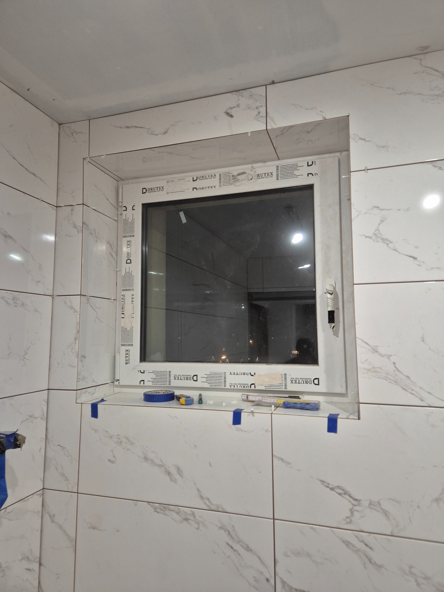 Białe płytki imitujące marmur wokół okna PCV w łazience. Widoczne narzędzia i niebieska taśma malarska. Etap wykończenia.