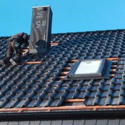 Remo Dach - Praca na dachu z czarnej dachówki: dekarz montuje komin i okno dachowe. Widoczne łatki i niebo w tle. Dachówka błyszczy, co sugeruje, że jest świeżo położona lub umyta.