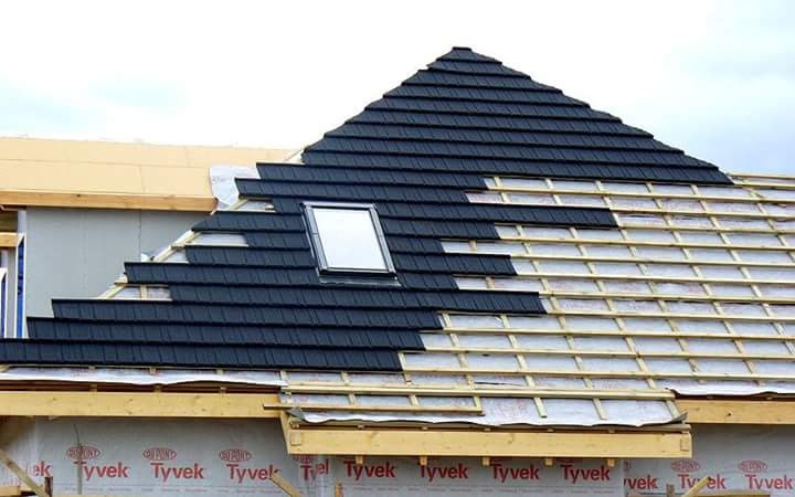 Fragment dachu w trakcie krycia, widoczne drewniane łaty, czarna dachówka układana rzędami i okno dachowe. Częściowo ukończony montaż.