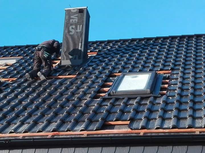 Praca na dachu z czarnej dachówki: dekarz montuje komin i okno dachowe. Widoczne łatki i niebo w tle. Dachówka błyszczy, co sugeruje, że jest świeżo położona lub umyta.