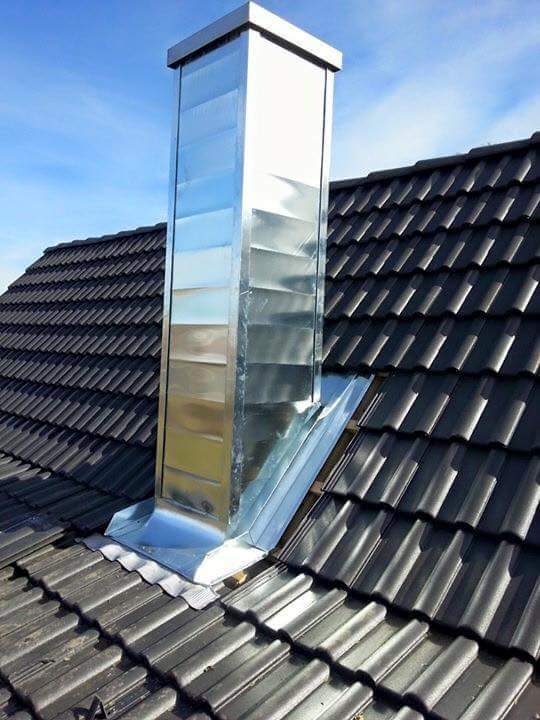 Błyszczący, metalowy komin wentylacyjny na dachu z ciemnej dachówki ceramicznej, zabezpieczony obróbką blacharską; widok z góry na tle błękitnego nieba.