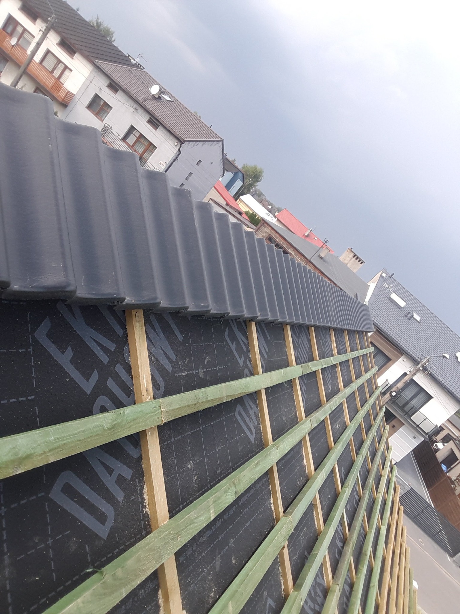 Fragment dachu w trakcie krycia: czarna membrana, drewniane łaty i rząd ułożonej szarej dachówki. W tle zabudowa mieszkalna.