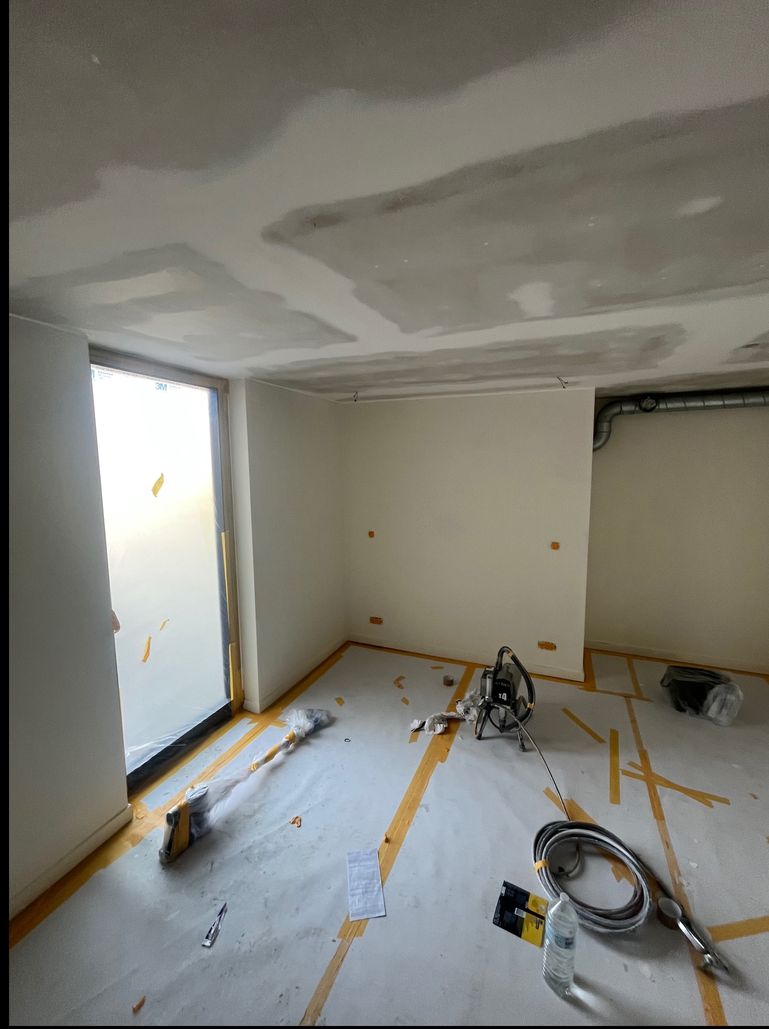 Pokój po szpachlowaniu, przygotowany do malowania. Okno zabezpieczone folią, na podłodze taśmy malarskie, widoczny agregat malarski i akcesoria.