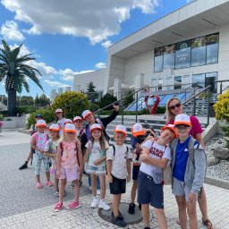 THE BEST CAMP - kolonie i obozy - Grupa dzieci w identycznych czapkach z opiekunką pozuje na tle budynku z napisem 'Restauracja i piętro'. Słoneczny dzień, wakacyjny klimat.