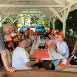 THE BEST CAMP - kolonie i obozy - Grupa nastolatków w pomarańczowych czapkach siedzi przy stole w altanie podczas obozu letniego. Widoczny opiekun. W tle drzewa i samochody.