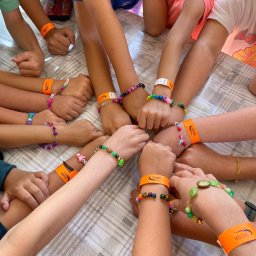 THE BEST CAMP - kolonie i obozy - Dziecięce dłonie z kolorowymi bransoletkami i pomarańczowymi opaskami 'DIUNA' na tle stołu w kratę, symbolizujące wspólną zabawę na kolonii lub obozie.