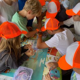 THE BEST CAMP - kolonie i obozy - Grupa dzieci w pomarańczowych czapkach tworzy biżuterię z koralików przy stole z kolorową grafiką, widoczne pudełka z akcesoriami i elementy dekoracyjne.