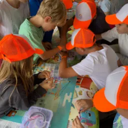 Grupa dzieci w pomarańczowych czapkach tworzy biżuterię z koralików przy stole z kolorową grafiką, widoczne pudełka z akcesoriami i elementy dekoracyjne.