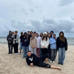 Grupa nastolatków i mężczyzna leżący na piasku plaży nad morzem, pochmurne niebo w tle. Zdjęcie z wycieczki lub obozu młodzieżowego.