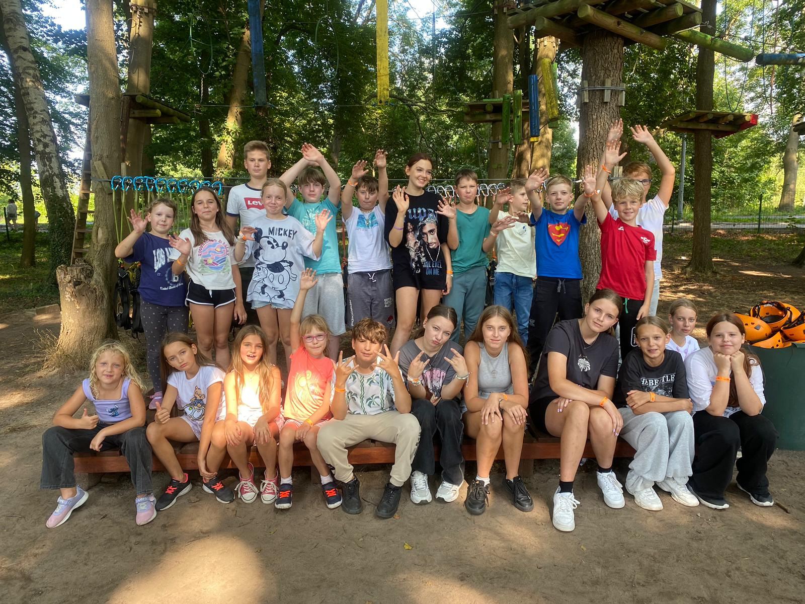 Grupa dzieci siedzi na ławce przed parkiem linowym, machając do kamery. W tle drzewa i elementy konstrukcji parku linowego.