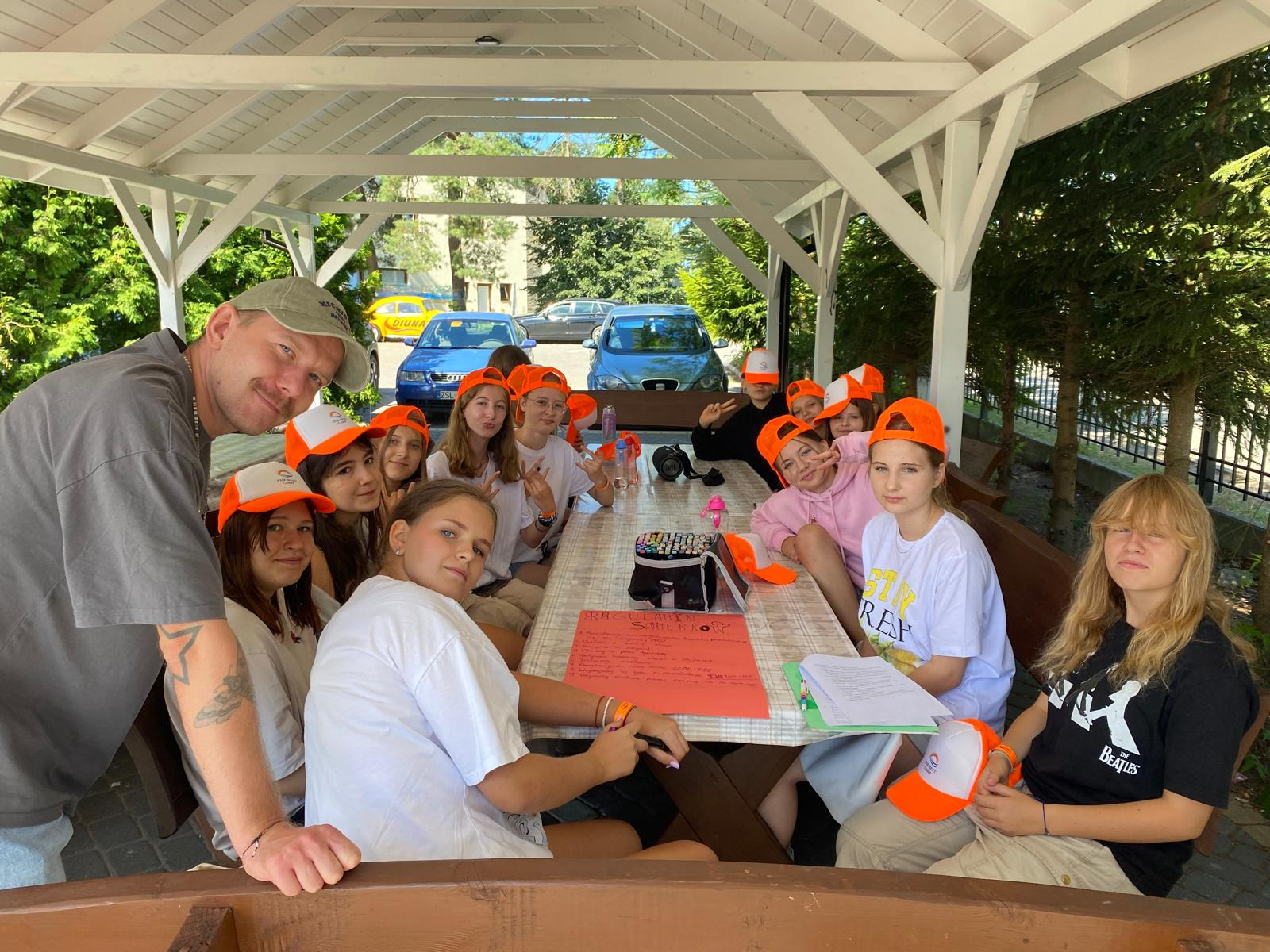 Grupa nastolatków w pomarańczowych czapkach siedzi przy stole w altanie podczas obozu letniego. Widoczny opiekun. W tle drzewa i samochody.
