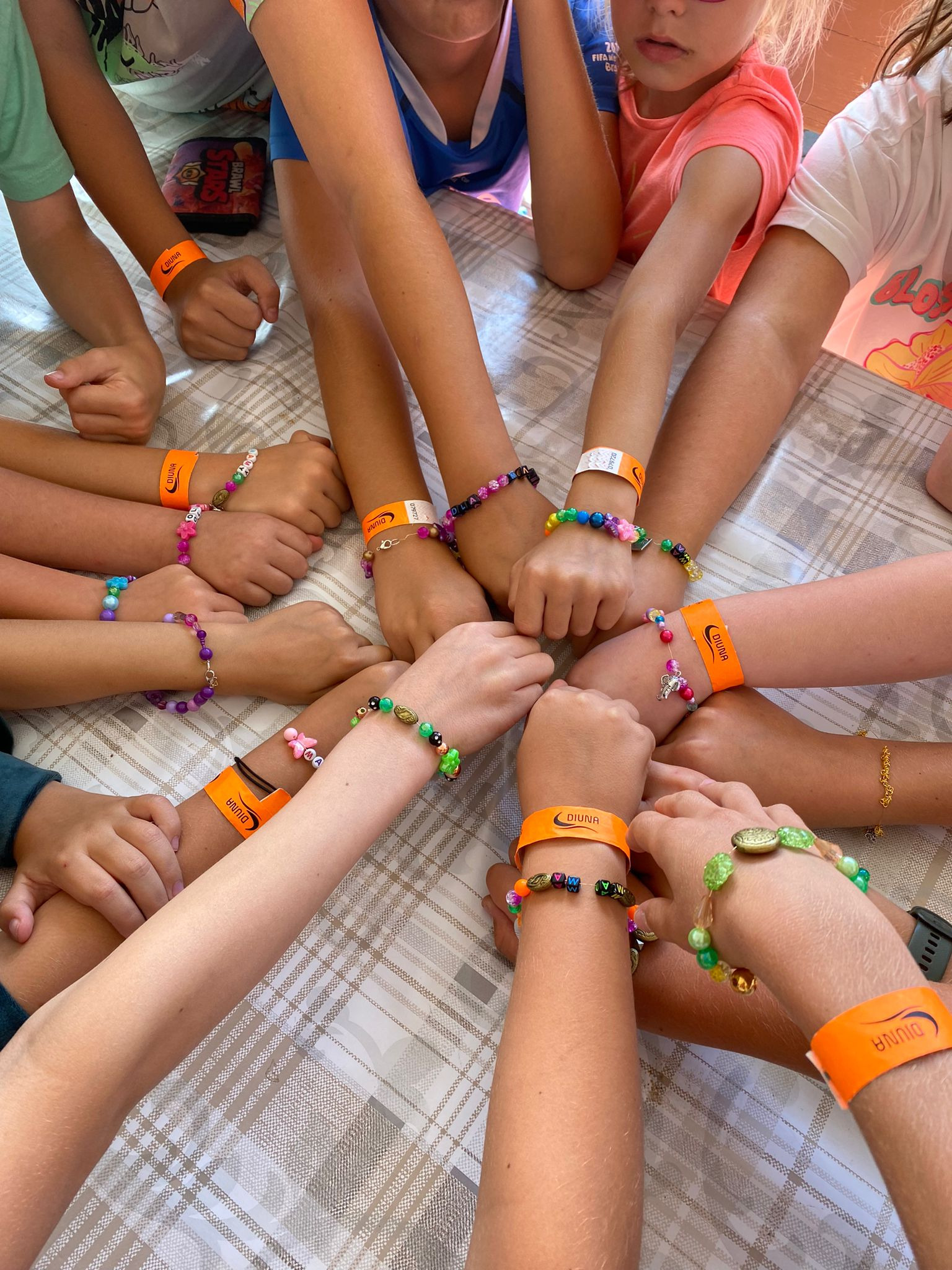 Dziecięce dłonie z kolorowymi bransoletkami i pomarańczowymi opaskami 'DIUNA' na tle stołu w kratę, symbolizujące wspólną zabawę na kolonii lub obozie.