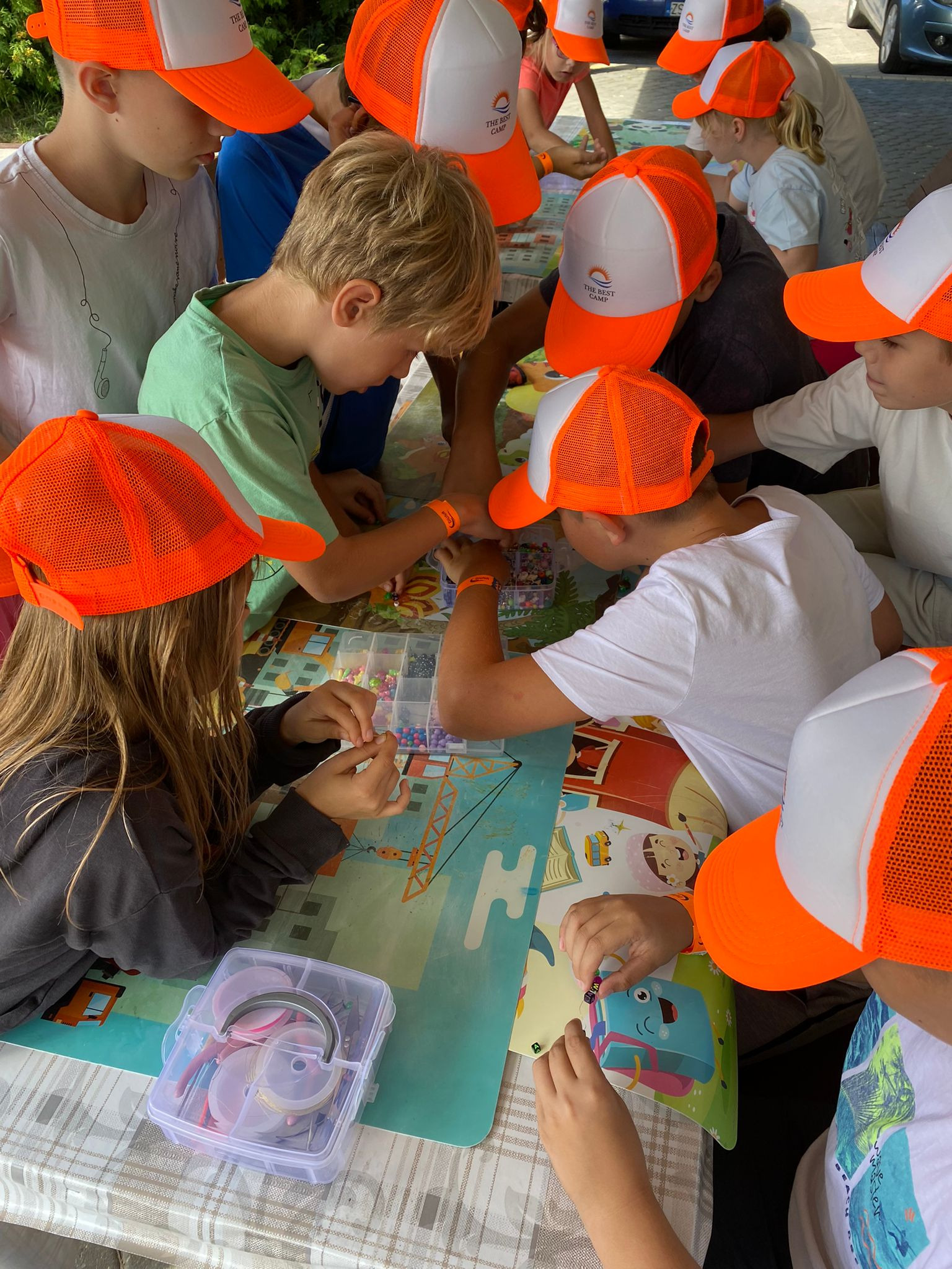 Grupa dzieci w pomarańczowych czapkach tworzy biżuterię z koralików przy stole z kolorową grafiką, widoczne pudełka z akcesoriami i elementy dekoracyjne.