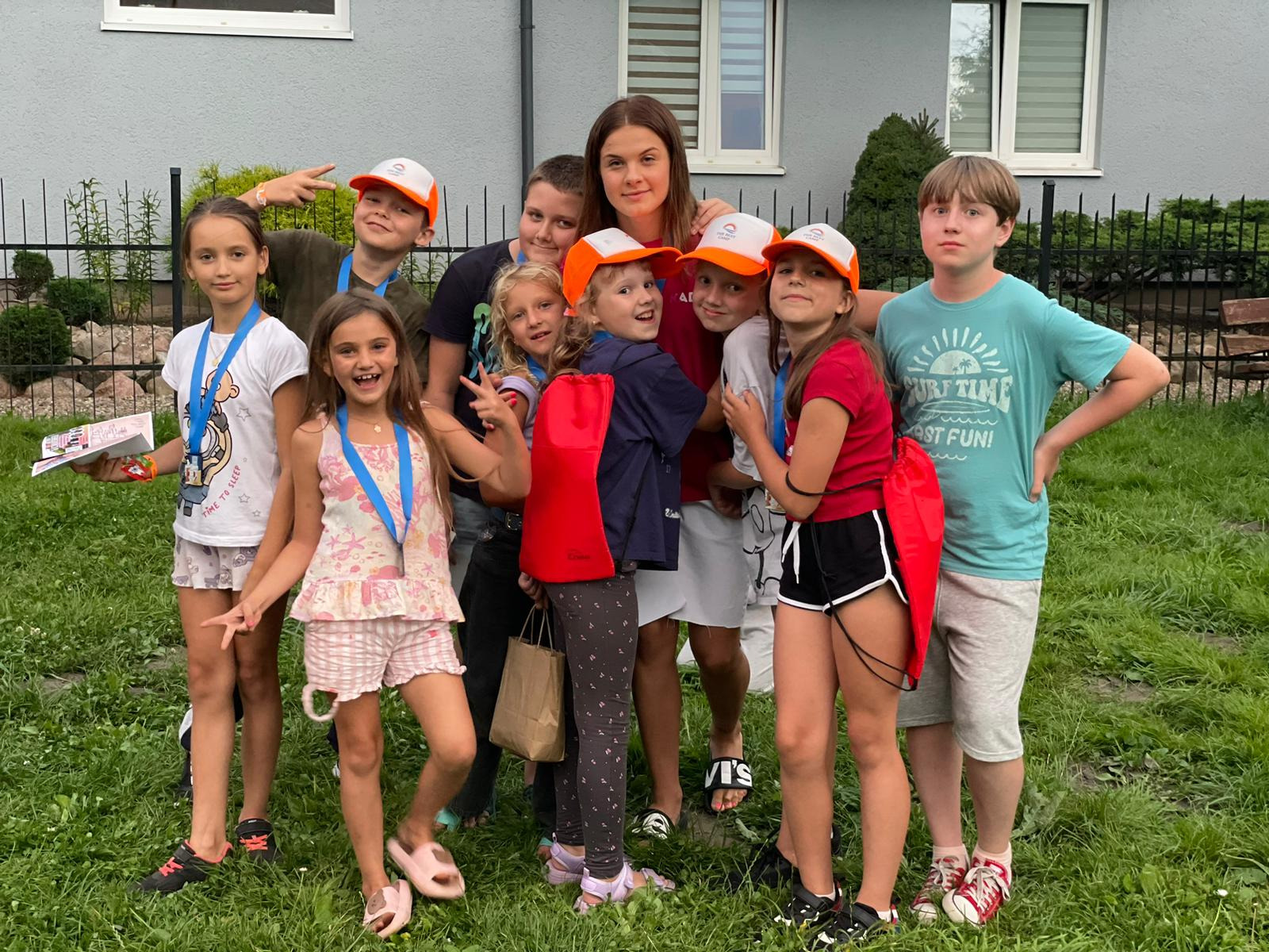 Grupa uśmiechniętych dzieci w czapkach i z medalami, pozujących na tle domu i zieleni, prawdopodobnie po zakończonych zajęciach lub wycieczce.