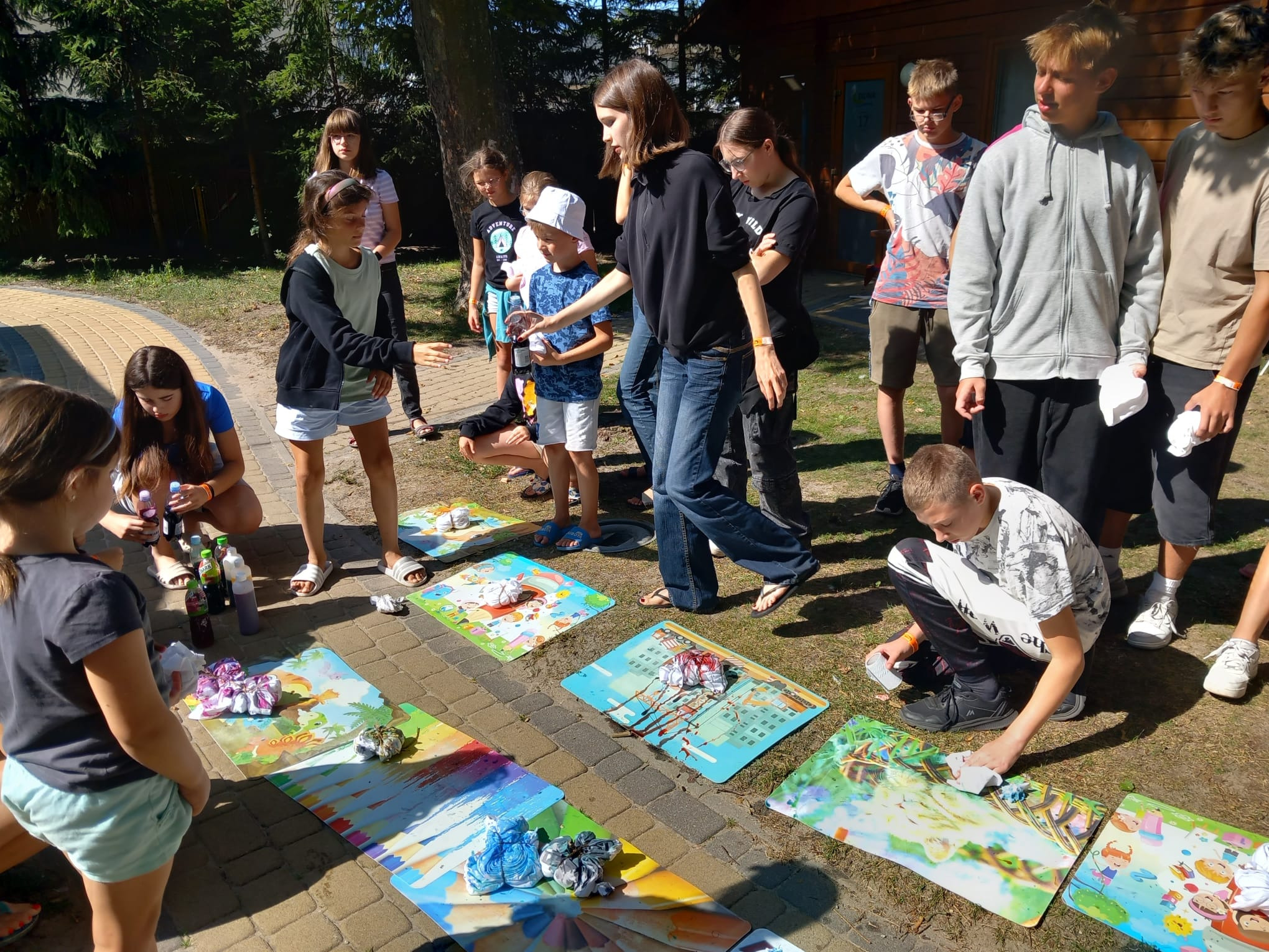 Grupa dzieci i dorosłych podczas warsztatów farbowania ubrań metodą tie-dye na świeżym powietrzu. Kolorowe maty z przygotowanymi koszulkami, butelki z barwnikami.