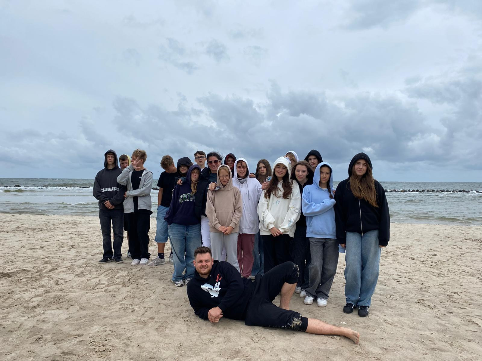 Grupa nastolatków i mężczyzna leżący na piasku plaży nad morzem, pochmurne niebo w tle. Zdjęcie z wycieczki lub obozu młodzieżowego.