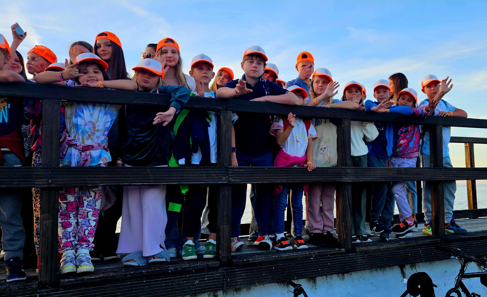 Grupa dzieci w pomarańczowych czapkach stoi na drewnianym pomoście, machając. W tle błękitne niebo i woda. Widok z perspektywy osoby stojącej poniżej pomostu.