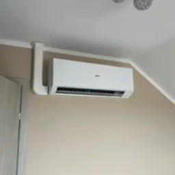 Biała klimatyzacja Haier zamontowana na ścianie w pokoju, z widoczną instalacją prowadzącą do sufitu. Na wyświetlaczu widoczna temperatura 20 stopni.