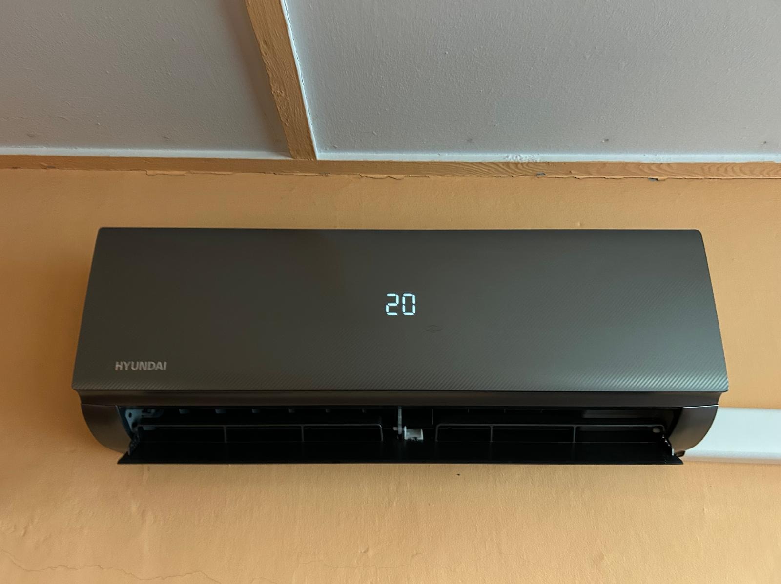 Szary klimatyzator Hyundai na pomarańczowej ścianie, wyświetlający temperaturę 20 stopni. Widoczne otwarte żaluzje. Ujęcie centralne z bliska.