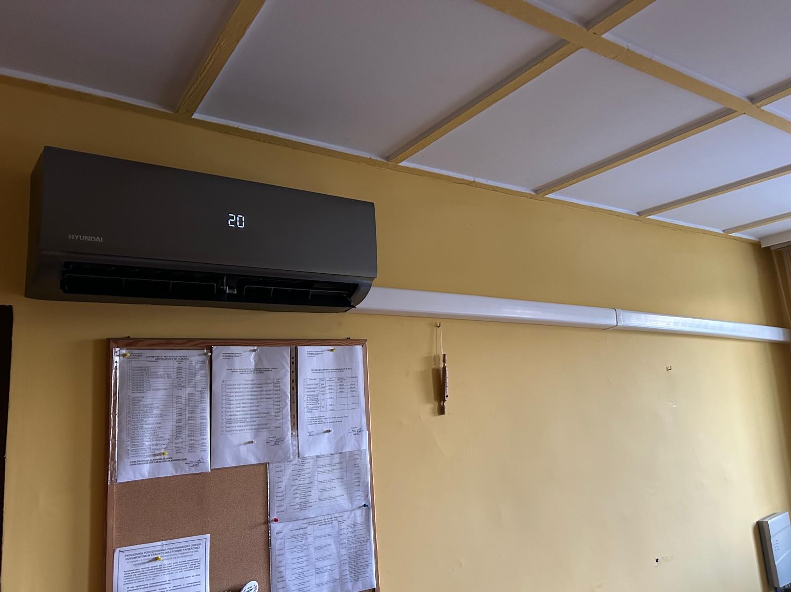 Szary klimatyzator Hyundai zamontowany na żółtej ścianie, z widocznymi przewodami i tablicą korkową z dokumentami. Widok z dołu, oświetlenie dzienne.