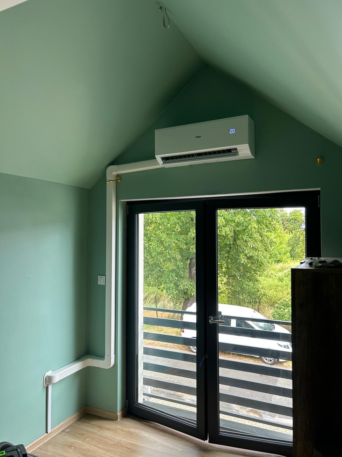 Klimatyzator Haier zamontowany w pokoju na poddaszu, z widocznymi przewodami w białej maskownicy. Za oknem widok na zieleń i zaparkowany samochód.