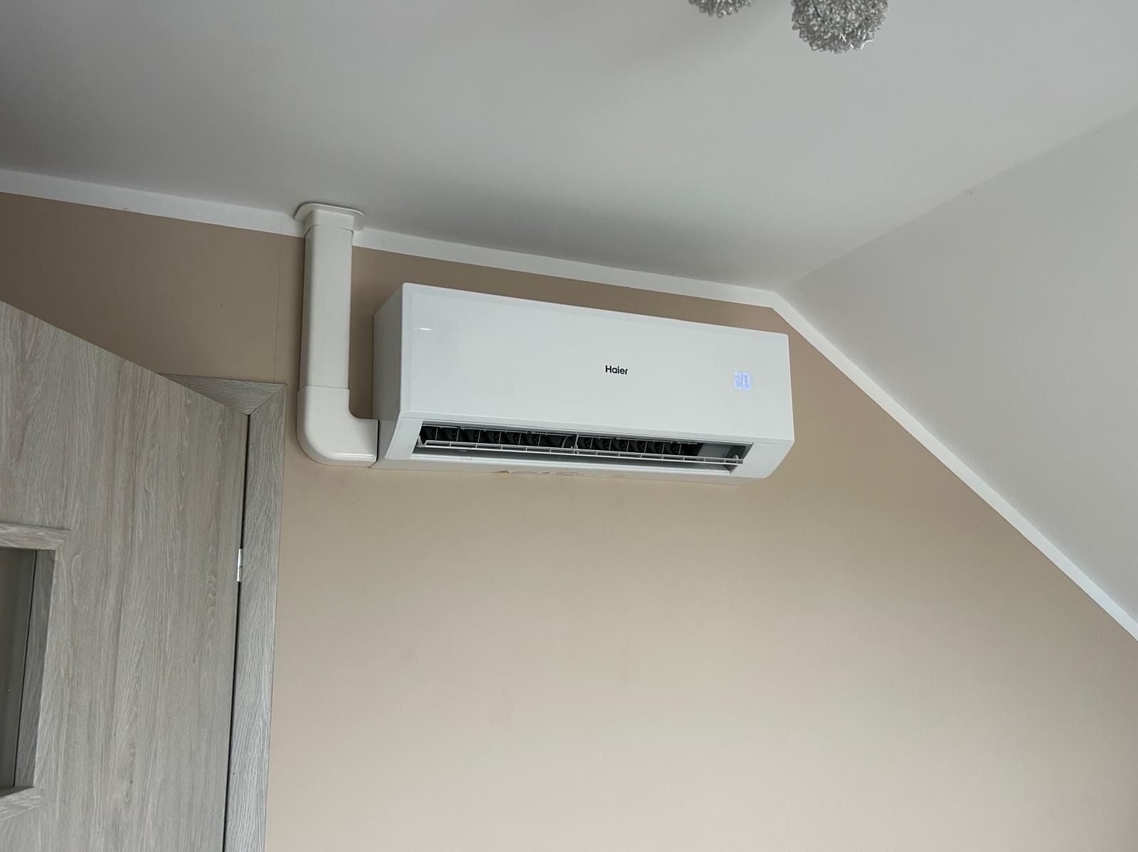 Biała klimatyzacja Haier zamontowana na ścianie w pokoju, z widoczną instalacją prowadzącą do sufitu. Na wyświetlaczu widoczna temperatura 20 stopni.