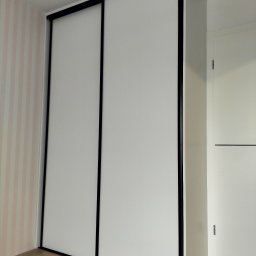 Pracownia Stolarska Hornet - Kamil Żeliszczak - Biała szafa przesuwna z czarnymi obramowaniami, wkomponowana w pokój z tapetą w pastelowe paski. Minimalistyczny design, widoczna podłoga z paneli.