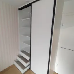 Biała szafa wnękowa z przesuwnymi drzwiami i czarną ramą, z wysuwanymi szufladami i półkami, na drewnianej podłodze, na tle ściany w pionowe, różowe paski.