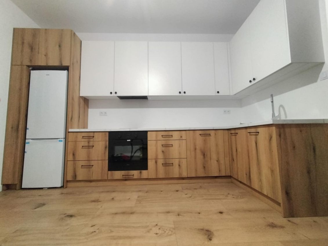 Nowoczesna kuchnia z białymi szafkami wiszącymi i dolnymi szafkami z drewnopodobnym fornirem. Uwagę zwraca zabudowana lodówka w pasującym stylu.