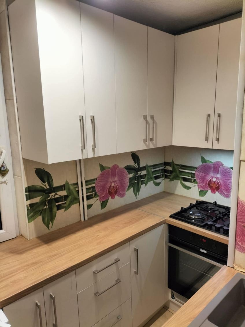 Jasna kuchnia z białymi szafkami i drewnianym blatem, ozdobiona motywem kwiatowym na ścianie. Widoczna płyta grzewcza i piekarnik. Minimalistyczny design.
