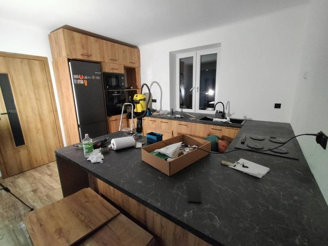 Montaż kuchni: blat z ciemnego marmuru, drewniane szafki, sprzęt AGD i narzędzia. Widoczny etap prac wykończeniowych, detale wnętrza.