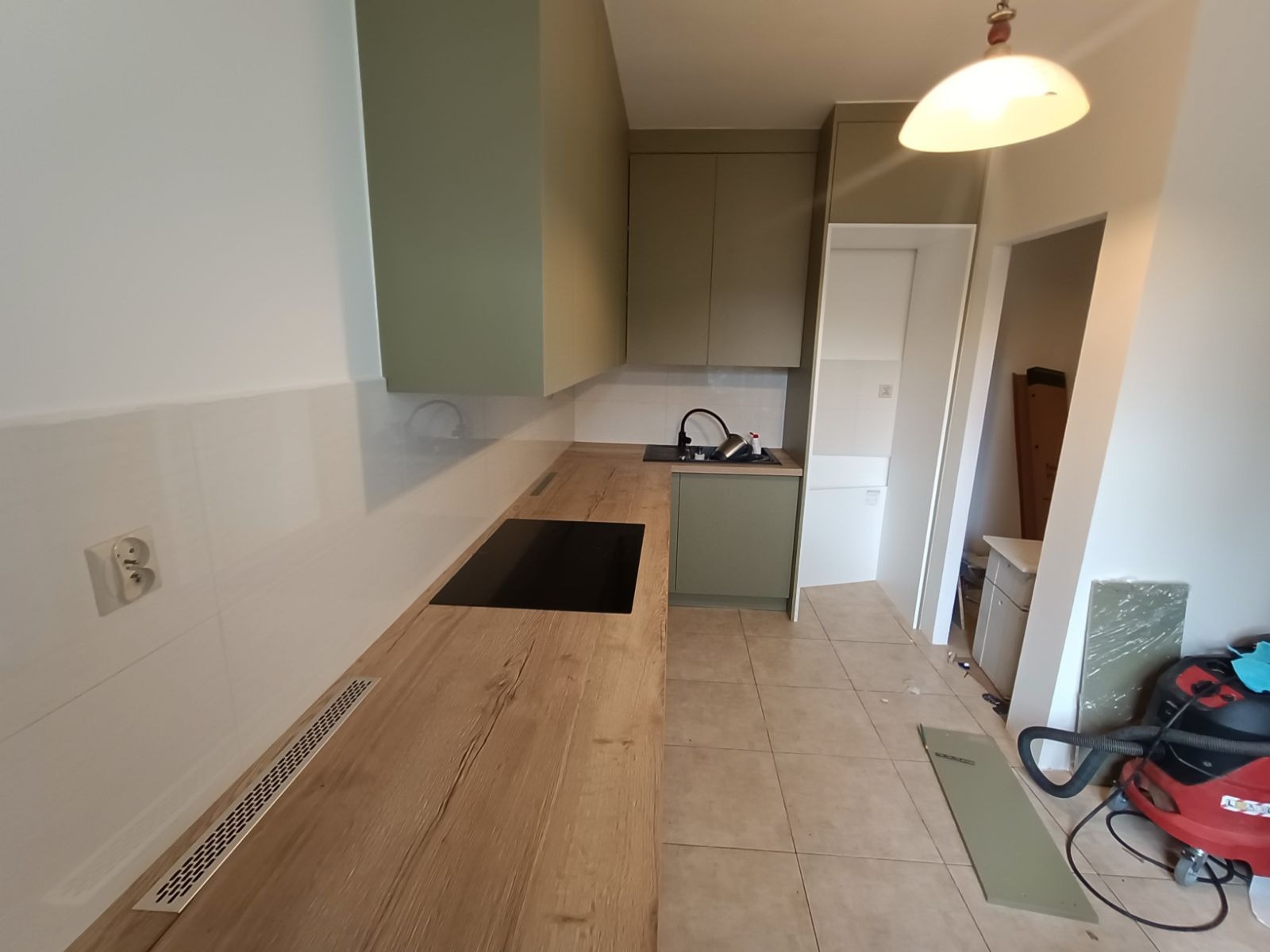 Kuchenna zabudowa w odcieniach zieleni, z drewnianym blatem i czarną płytą. Widoczny fragment podłogi z płytek i sprzęt sprzątający. Ujęcie z perspektywy narożnika.