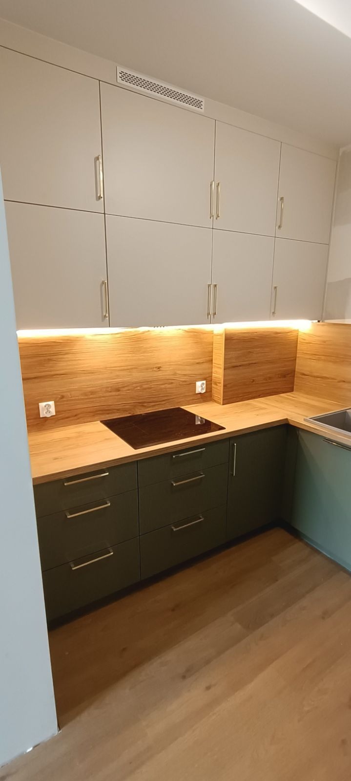 Nowoczesna kuchnia z szarymi szafkami górnymi i oliwkowymi dolnymi, drewnianym blatem i oświetleniem LED pod szafkami. Widoczna płyta indukcyjna i drewniana podłoga.