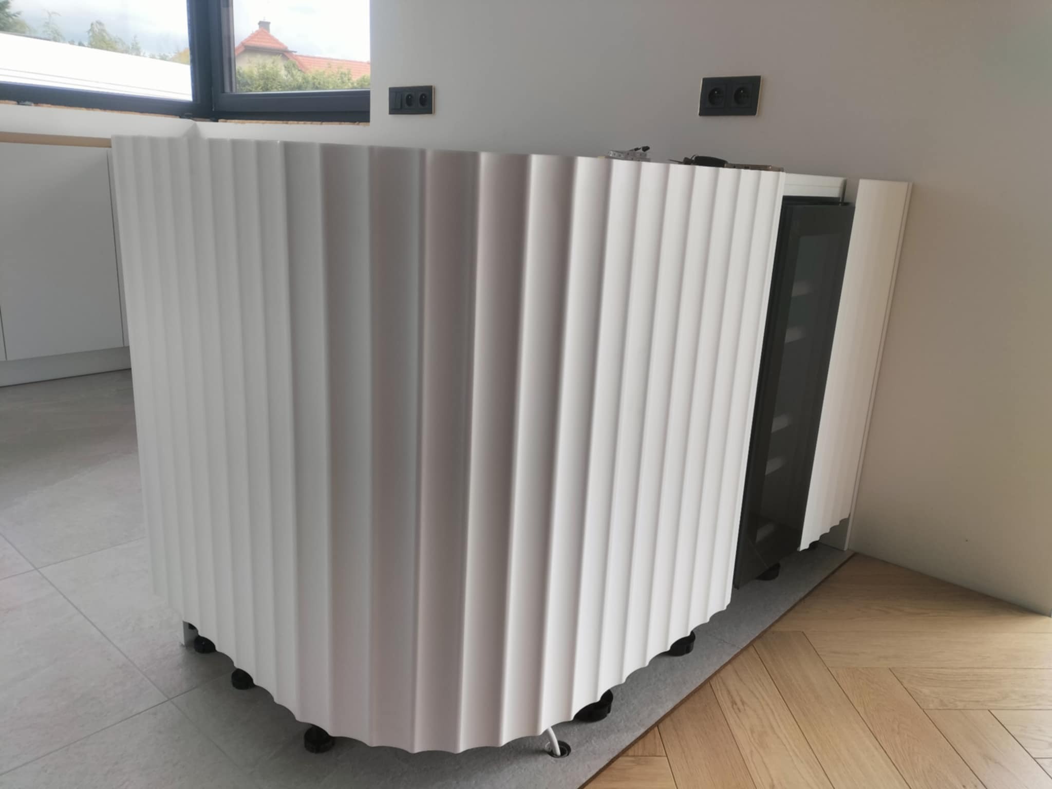 Nowoczesna, biała zabudowa barkowa z ryflowanym frontem, na tle jasnej ściany z gniazdkami. Widoczny fragment podłogi z płytek i parkietu.