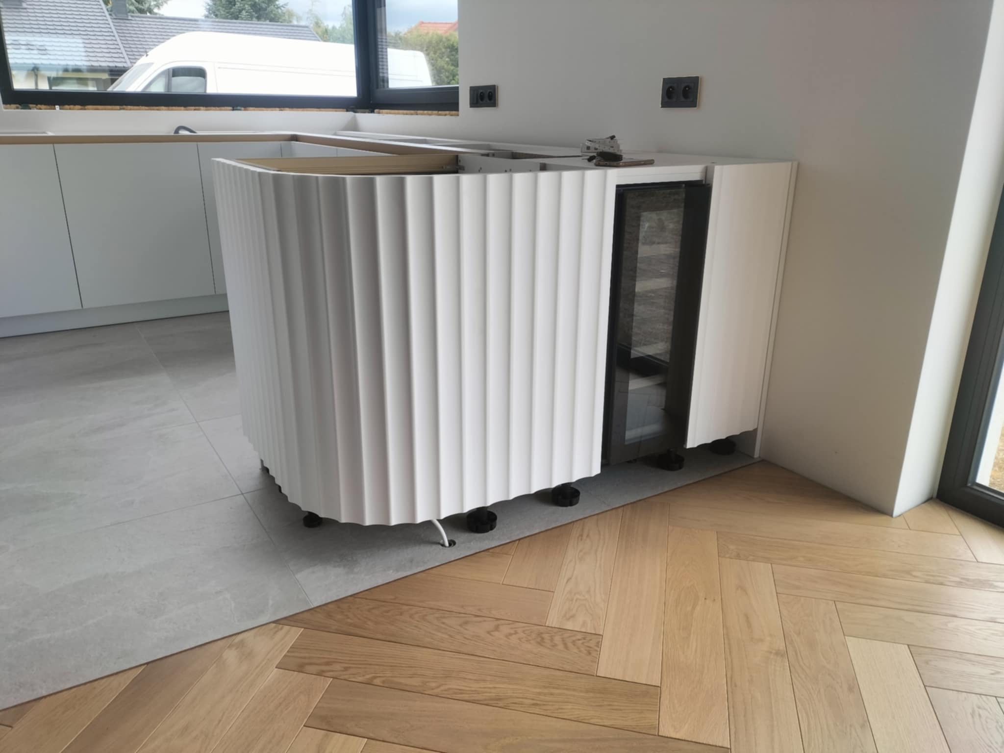 Fragment montażu białej, falistej zabudowy wyspy kuchennej z wbudowaną chłodziarką do wina. Widoczne regulowane nóżki i fragment podłogi z parkietu w jodełkę.