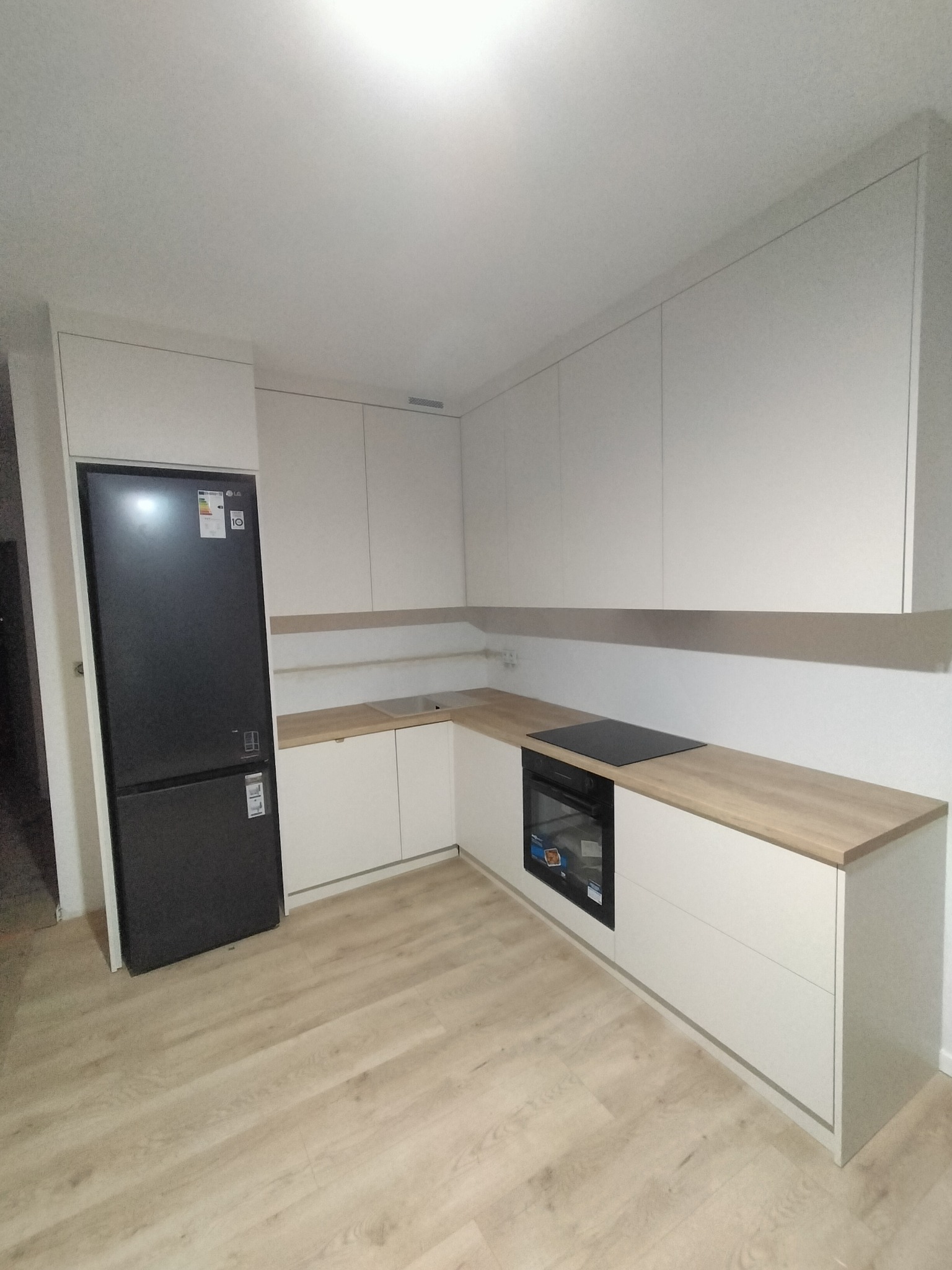 Jasna kuchnia z drewnianym blatem, czarną lodówką i płytą indukcyjną. Minimalistyczny design, proste fronty szafek, nowoczesne oświetlenie. Widok na całą zabudowę kuchenną.