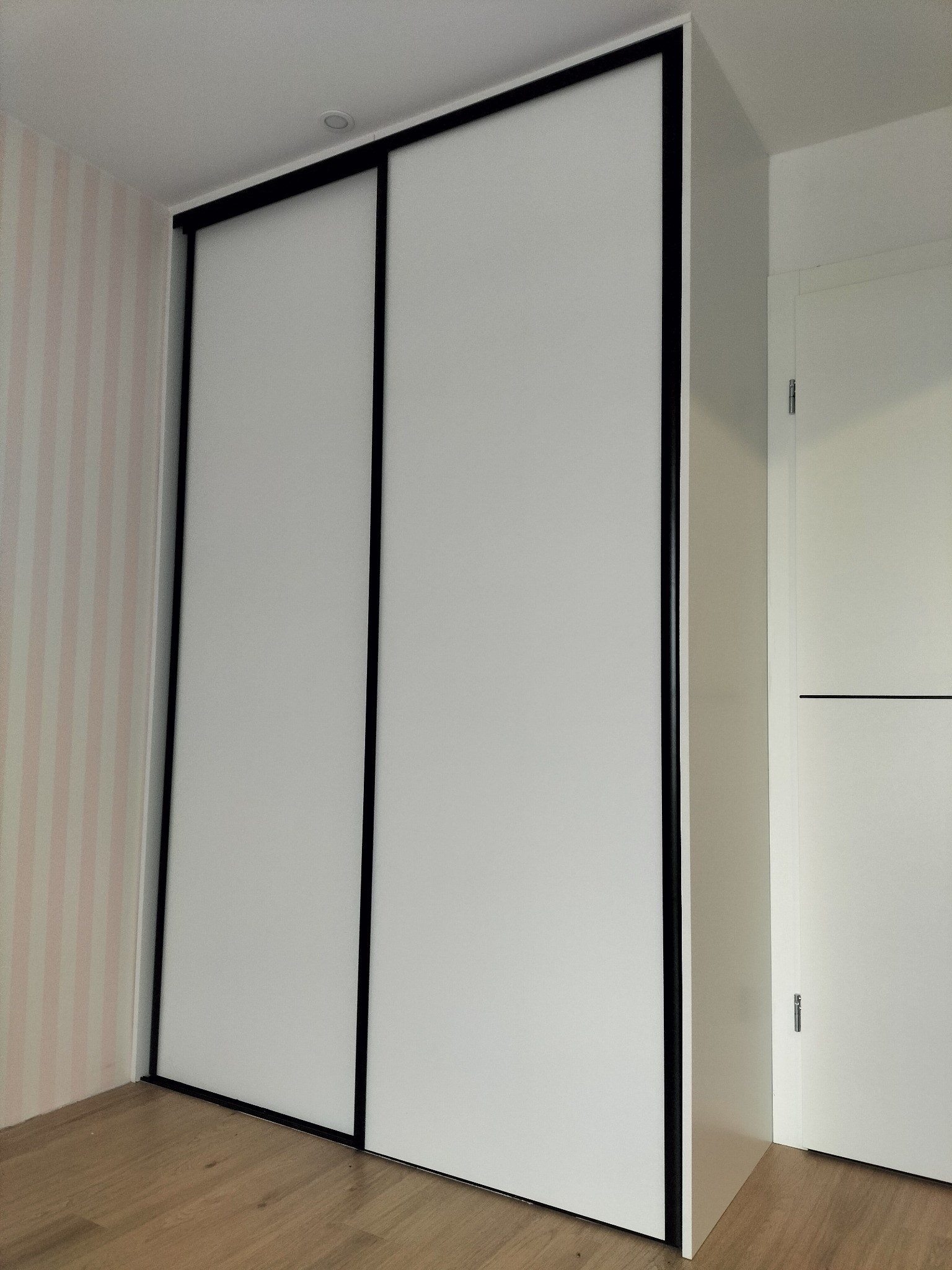 Biała szafa przesuwna z czarnymi obramowaniami, wkomponowana w pokój z tapetą w pastelowe paski. Minimalistyczny design, widoczna podłoga z paneli.