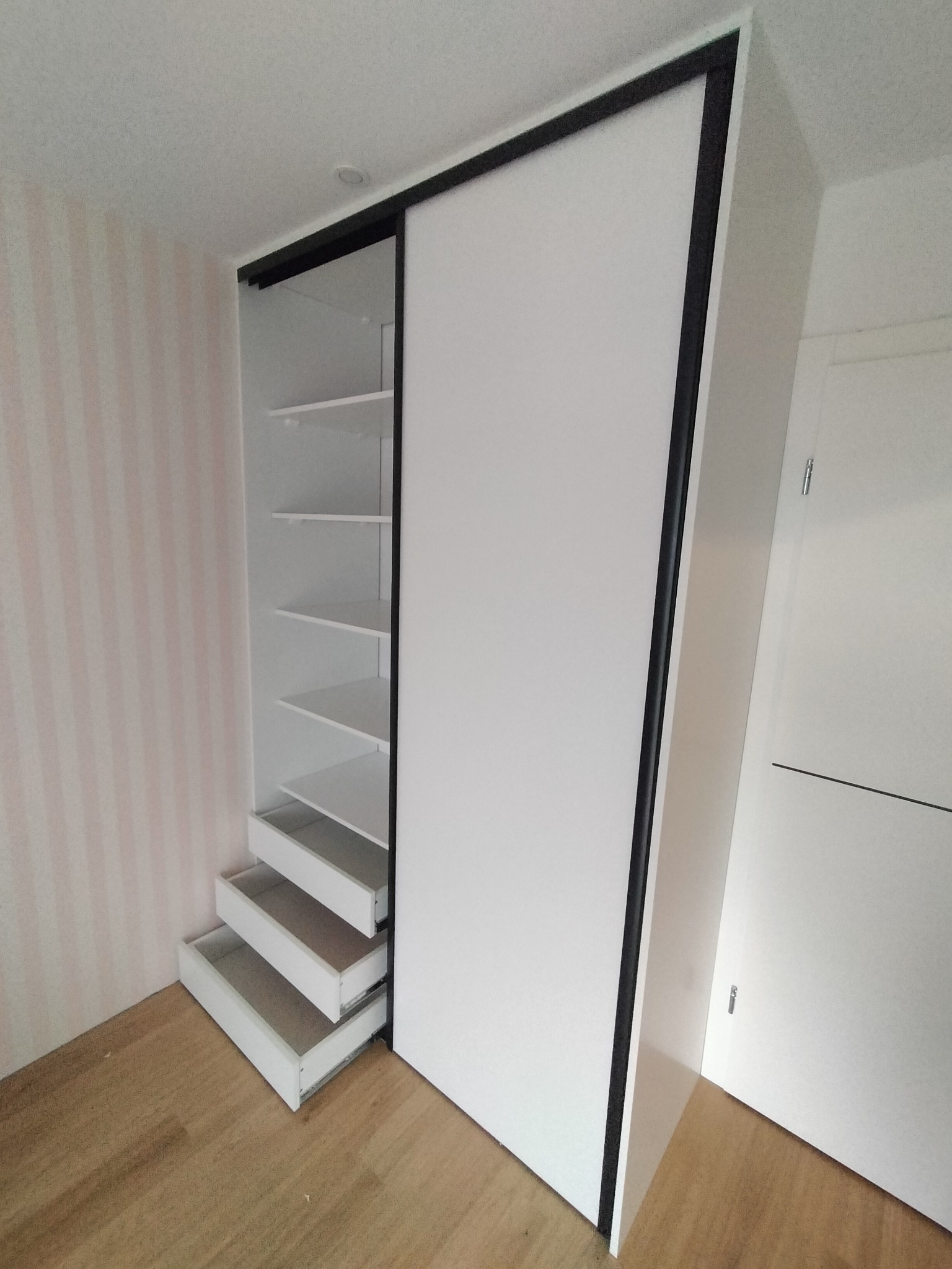 Biała szafa wnękowa z przesuwnymi drzwiami i czarną ramą, z wysuwanymi szufladami i półkami, na drewnianej podłodze, na tle ściany w pionowe, różowe paski.