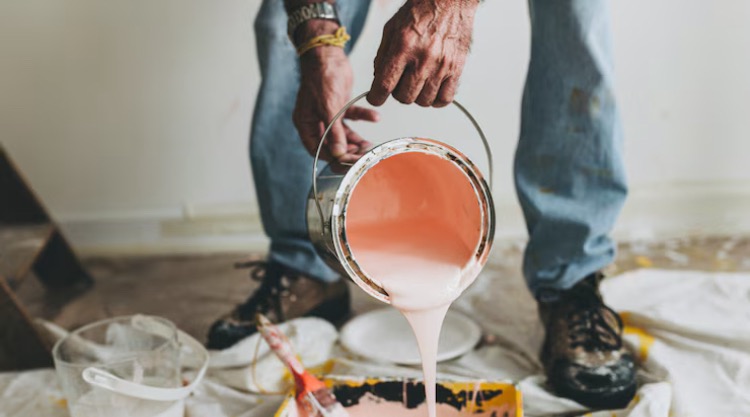 Osoba w jeansach wylewa różową farbę z puszki do kuwety malarskiej na zabezpieczonej folią podłodze. Widoczne narzędzia i fragment pomalowanej ściany.