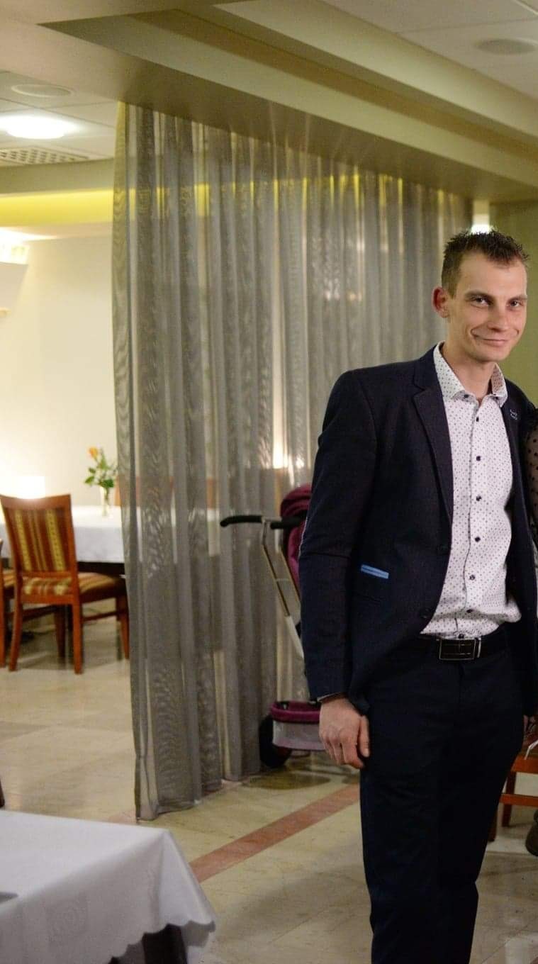 Mężczyzna w garniturze stoi w jasnym wnętrzu z zasłonami, obok nakrytego stołu i wózka dziecięcego, spoglądając w obiektyw z uśmiechem.