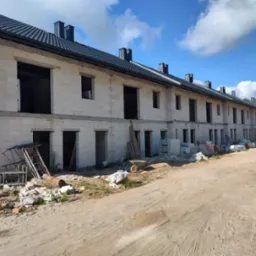 Wznoszenie szeregu dom&oacute;w z białych bloczk&oacute;w, z widocznymi otworami okiennymi i drzwiowymi. Na pierwszym planie materiały budowlane i narzędzia.