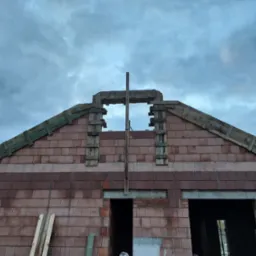 Wznoszenie domu z keramzytu: widok na fragment ściany szczytowej z otworami okiennymi w trakcie murowania, z drewnianym szalunkiem i pochmurnym niebem w tle.